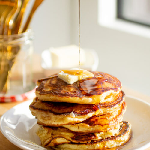 A stack of golden pancakes on a white plate topped with butter and syrup.