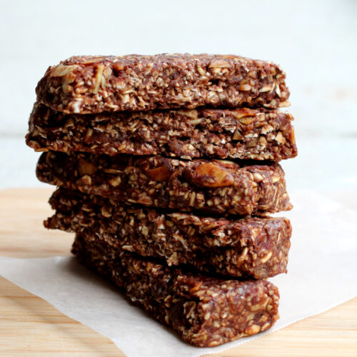Five date-sweetened granola bars stacked on a sheet of parchment paper on a cutting board.