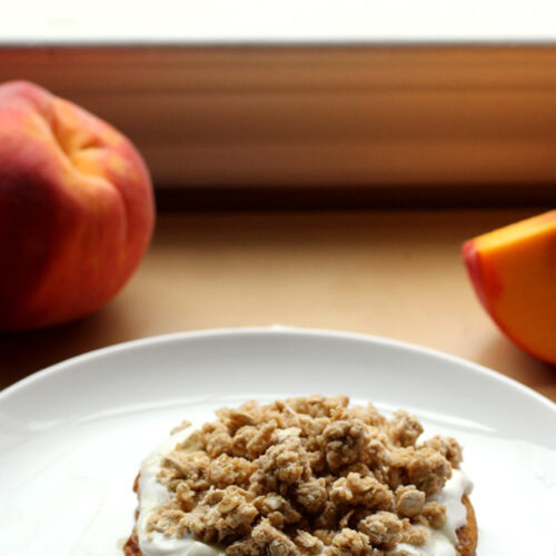 A stack of pancakes on a white plate layered with yogurt, peach slices and granola with peaches in the background.