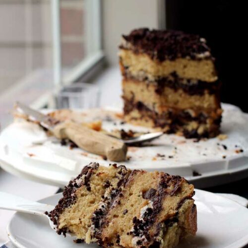 A slice of a triple layer chocolate chip cake layered with chocolate crumbs on a white plate with the rest of the cake in the background.