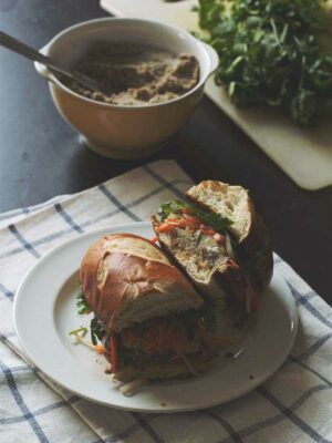 A banh mi cut in half on a white plate to show the shredded veggies and tofu in the middle.