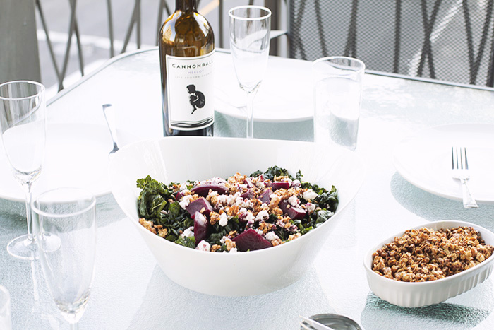 white bowl filled with kale, goat cheese, beets and walnut granola on a glass table with wine glasses and other plates