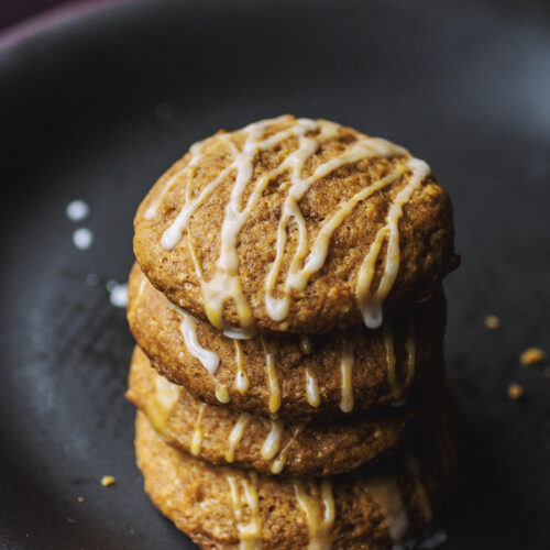 A stack of four pumpkin cookies drizzled with a white glaze on a black plate.