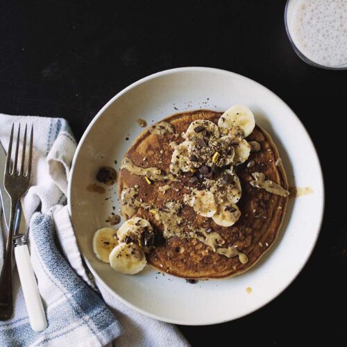 A golden brown pancake topped with sliced banana and peanut butter on a white plate next to a fork laid on top of a white towel.