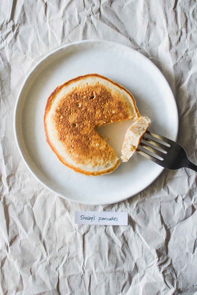 A pancake with a bite taken out of it with a fork on a white plate with the label \"Shelby\'s Pancakes.\"