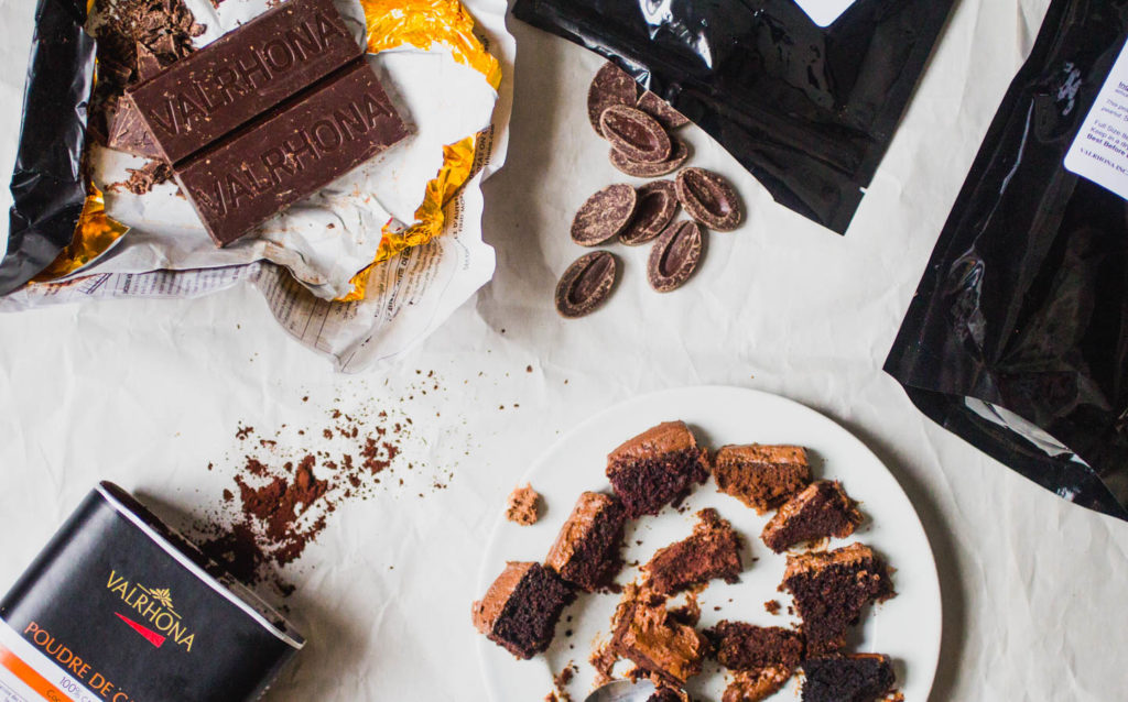 Half eaten slices of cake on a white plate next to a Valrhona chocolate bar, chocolate feves, and cocoa powder.
