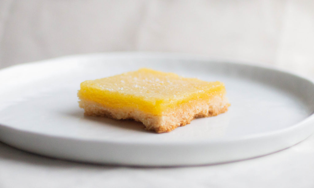 single slice of mel's kitchen cafe lemon bars on white plate featuring equal ratio of crust and lemon curd