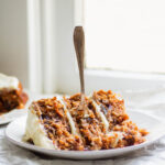 a slice of carrot cake with a fork on a white plate next to a window.