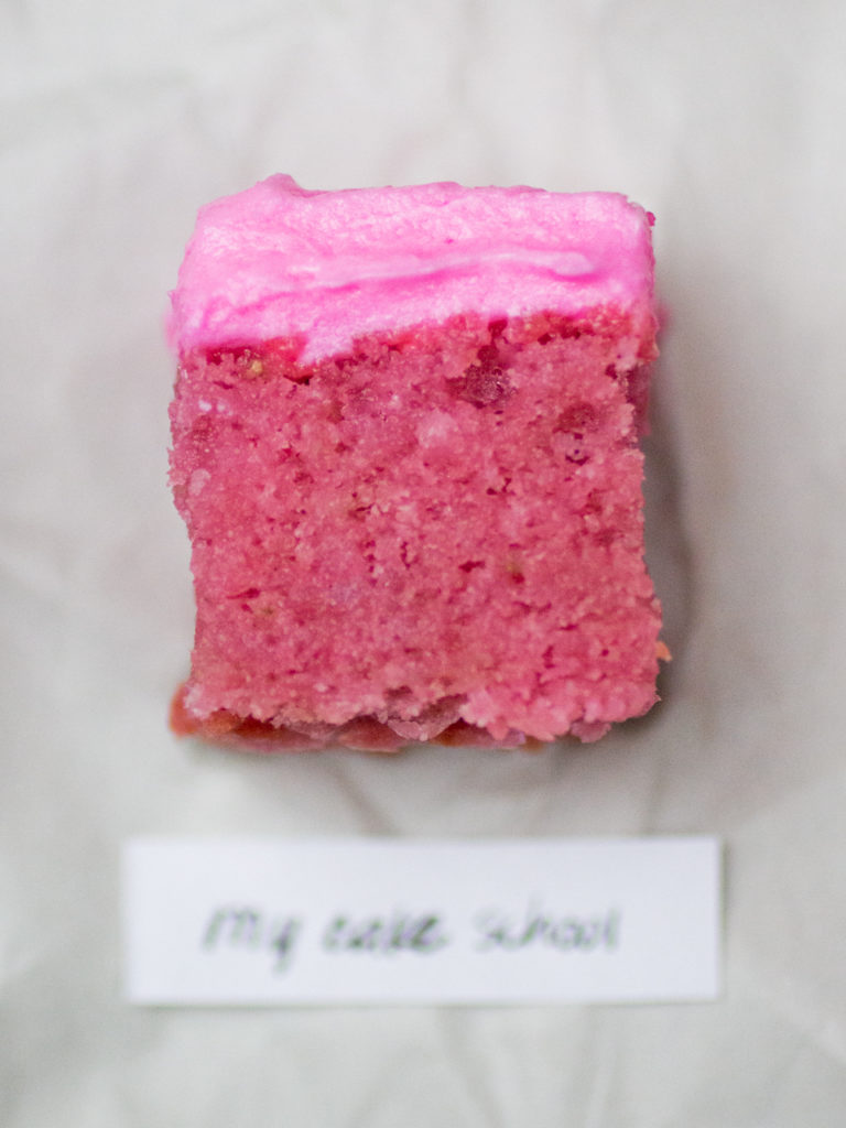 Single rectangular slice of bright pink iced strawberry cake by My Cake School on white parchment paper