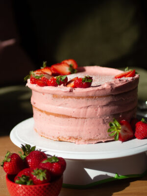 strawberry cake on a white cake stand with a bowl of strawberries.