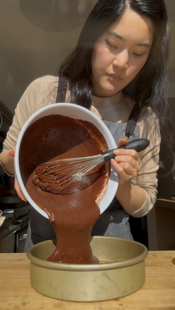 A girl pouring a white bowl of chocolate cake batter into an 8" cake pan.
