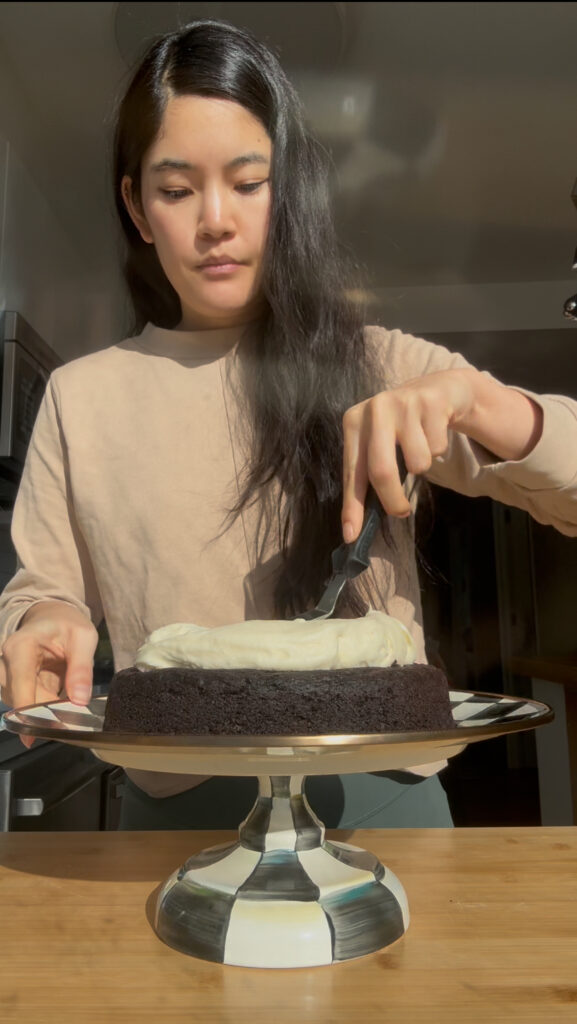 A girl in a brown sweater frosting a chocolate cake with cream cheese frosting.