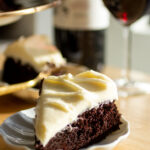A slice of chocolate cake with cream cheese frosting on a white plate with a glass of red wine in the background.