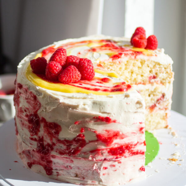 A three layer vanilla cake with raspberries and lemon curd on a cake stand with a slice cut out to show the interior.