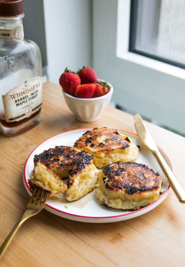 Trois pancakes épais sur une assiette blanche avec un bord rouge à côté d'un bol de fraises et d'un pot de sirop d'érable.