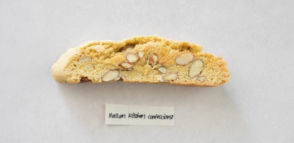 A pice of biscotti on a gray background with the label "Italian Kitchen Confessions."