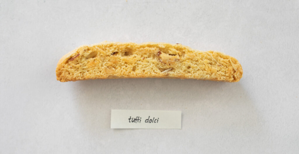 A pice of biscotti on a gray background with the label "Tutti Dolci."