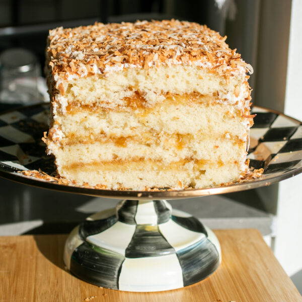 A four-layer coconut cake covered in toasted coconut on a black and white cake stand with a slice taken out to show the cross-section.