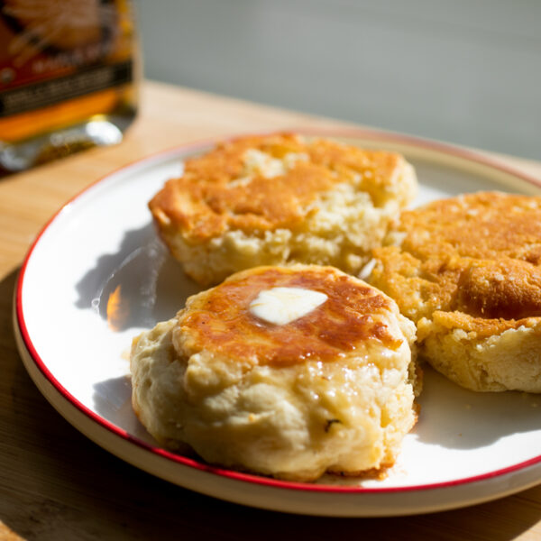 Three thick pancakes with golden surfaces on a white plate with maple syrup in the background.